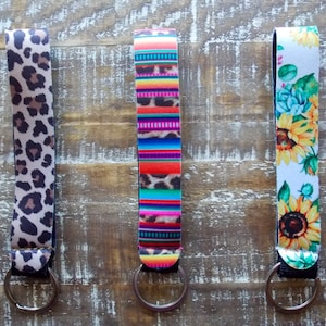 May include: Three keychains with different patterns. The first keychain has a leopard print pattern. The second keychain has a colorful striped pattern. The third keychain has a floral pattern with sunflowers.