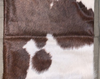 May include: A brown and white cowhide pillow with a soft, plush texture. The pillow is rectangular and has a subtle sheen.