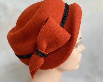 May include: A burnt orange felt hat with a large bow and black trim. The hat is styled with a folded brim and a decorative band around the crown. The bow is positioned on the side, adding a touch of elegance.
