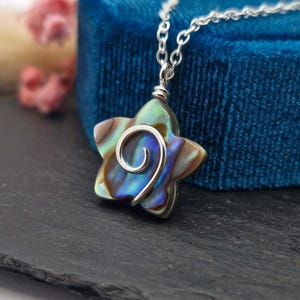 May include: A star-shaped pendant necklace with a silver chain. The pendant has a swirling silver design over a colourful abalone shell. The shell displays iridescent blues, greens, and browns. The necklace rests on a dark grey surface with a blue velvet box in the background.