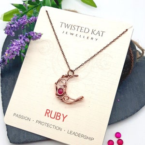 May include: Copper wire crescent moon necklace with a ruby gemstone. The necklace is on a card that says "Twisted Kat Jewellery" and "Ruby Passion Protection Leadership".
