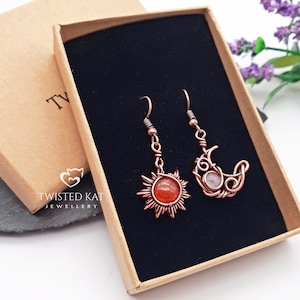 May include: A pair of copper wire earrings with gemstone accents. One earring features a sun design with a red-orange stone, and the other earring features a crescent moon design with a white stone.