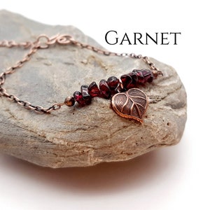 May include: A copper bracelet with a row of dark red garnet stones and a leaf charm. The bracelet is displayed on a grey stone, with the word "GARNET" in white text.