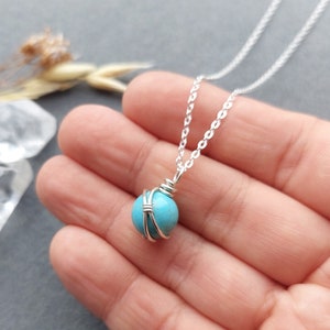 May include: A silver chain necklace with a turquoise gemstone wrapped in silver wire. The necklace is on a hand with a light grey background.