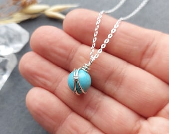 May include: A silver chain necklace with a turquoise gemstone wrapped in silver wire. The necklace is on a hand with a light grey background.