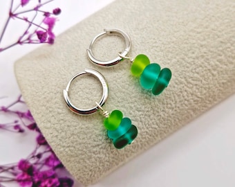May include: A pair of silver hoop earrings featuring dangling charms made of stacked, frosted sea glass beads in shades of lime green and teal. Each earring consists of a circular silver hoop with three small, rounded glass beads attached.