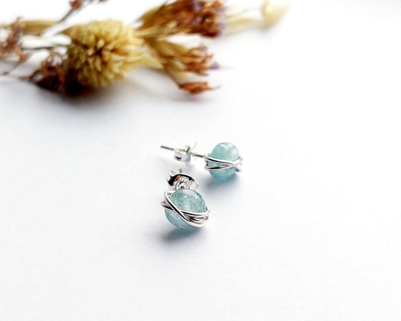 May include: A pair of stud earrings featuring light blue, spherical gemstones wrapped in silver wire. The earrings are set against a white background, with dried flowers in the upper left corner. The earrings are small and delicate.