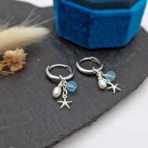以下が含まれることがあります： シルバーのフープピアス。各イヤリングには、白い真珠、水色のビーズ、小さなヒトデのチャームが付いています。イヤリングは濃い灰色の表面に表示され、背景には青いベルベットのジュエリーボックスがあります。