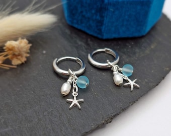 May include: Silver hoop earrings with dangling charms. Each earring features a white pearl, a light blue bead, and a small starfish charm. The earrings are displayed on a dark gray surface, with a blue velvet jewelry box in the background.