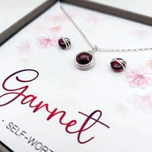 K&ouml;nnte beinhalten: Ein silbernes Halsketten- und Ohrring-Set mit einem roten Granatsteine. Die Halskette hat eine zarte Kette und die Ohrringe sind klein und schlicht. Das Set ist in einer wei&szlig;en Schachtel mit dem Wort "Granat" in einem rosafarbenen Farbverlauf verpackt.