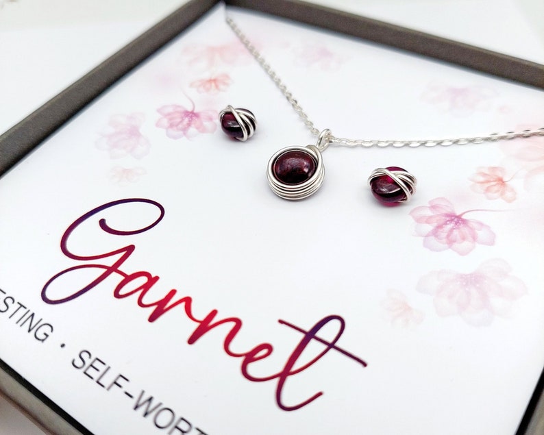 K&ouml;nnte beinhalten: Ein silbernes Halsketten- und Ohrring-Set mit einem roten Granatsteine. Die Halskette hat eine zarte Kette und die Ohrringe sind klein und schlicht. Das Set ist in einer wei&szlig;en Schachtel mit dem Wort "Granat" in einem rosafarbenen Farbverlauf verpackt.