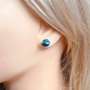 May include: A pair of small, teal blue gemstone earrings with silver wire wrapping. The earrings are stud style and are shown on a model's ear.