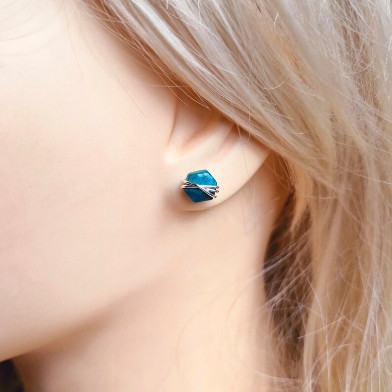 May include: A pair of small, teal blue gemstone earrings with silver wire wrapping. The earrings are stud style and are shown on a model's ear.