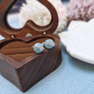 May include: A pair of aquamarine stud earrings with a silver wire design, displayed in a heart-shaped wooden jewelry box. The earrings have a light blue, spherical gemstone. The box is open, revealing a brown interior. A white decorative plate is in the background.