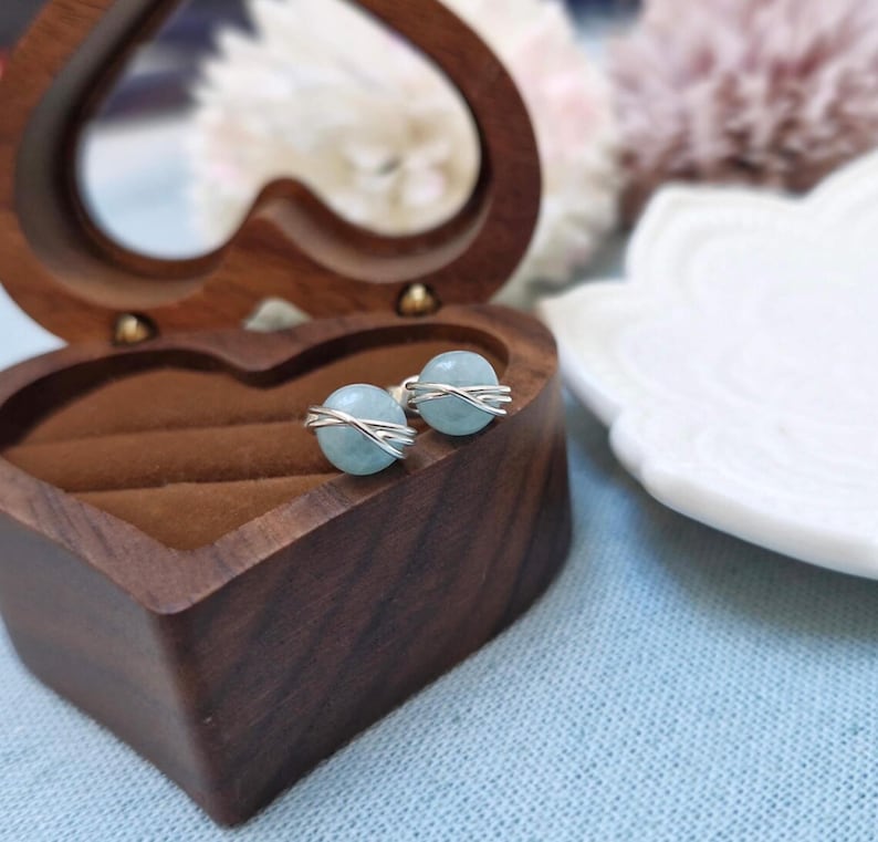 May include: A pair of aquamarine stud earrings with a silver wire design, displayed in a heart-shaped wooden jewelry box. The earrings have a light blue, spherical gemstone. The box is open, revealing a brown interior. A white decorative plate is in the background.