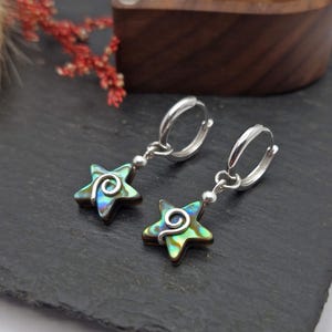 May include: Silver hoop earrings with star-shaped pendants. The pendants have a swirling silver design over a green and blue iridescent shell. The earrings are presented on a dark grey surface.