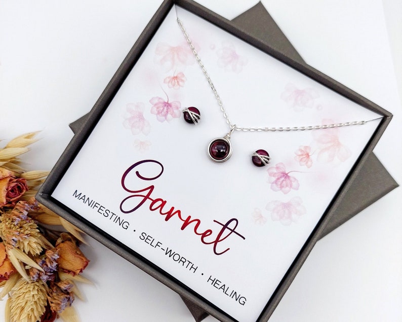 K&ouml;nnte beinhalten: Ein silbernes Halsketten- und Ohrring-Set mit roten Granatsteinen in einer wei&szlig;en Schachtel mit den Worten "Granat, Manifestieren, Selbstwertgef&uuml;hl, Heilung" darauf.