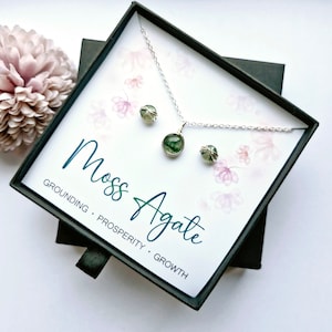 May include: A moss agate necklace and earrings set in a black gift box. The jewelry is silver and features green moss agate stones. The box has a white background with the words "Moss Agate" and "Grounding, Prosperity, Growth" printed on it.