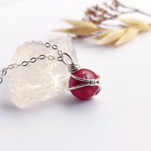 May include: A silver necklace with a magenta gemstone pendant. The round gemstone is held in place by a wire wrap, and hangs from a fine silver chain. The background is white, with a clear crystal and dried plants. The pendant is approximately 1.5 cm in diameter.