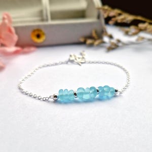 May include: A delicate silver chain bracelet with a row of light blue sea glass beads. The bracelet is on a white surface.