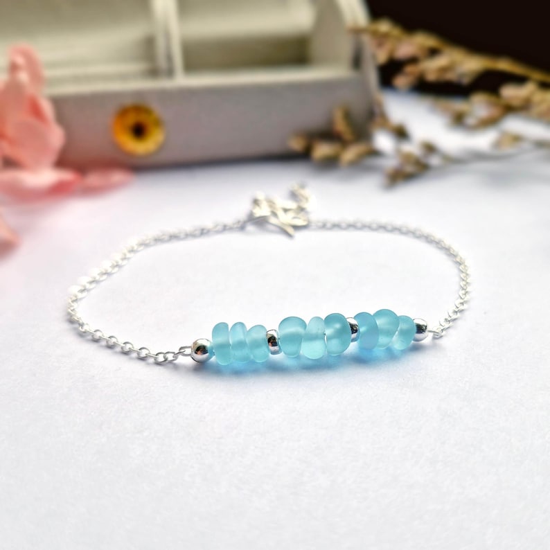 May include: A delicate silver chain bracelet with a row of light blue sea glass beads. The bracelet is on a white surface.