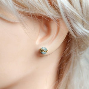 May include: A close-up of a light blue gemstone stud earring with gold wire wrapping. The earring is set against the backdrop of a person's ear and skin, with blonde hair visible in the background. The earring is a small, round shape.