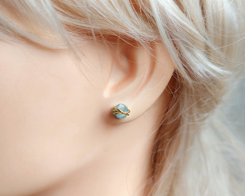 May include: A close-up of a light blue gemstone stud earring with gold wire wrapping. The earring is set against the backdrop of a person's ear and skin, with blonde hair visible in the background. The earring is a small, round shape.