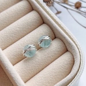 May include: A pair of round, light blue gemstone stud earrings. Each earring features a polished, spherical stone wrapped with a silver-colored wire. The earrings are displayed inside a beige jewelry box.