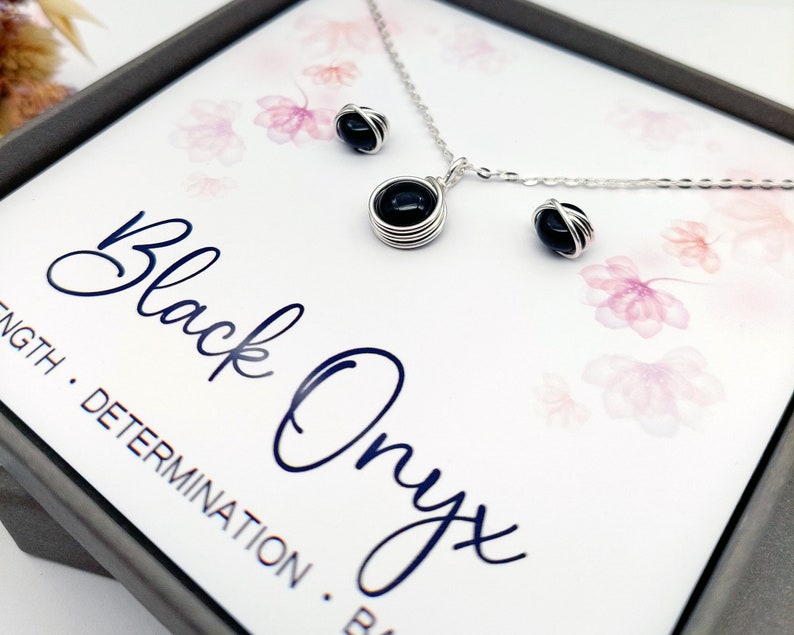 May include: A black onyx necklace and earrings set in a silver wire wrapped design. The jewelry is displayed in a box with the words "Black Onyx" and "Strength. Determination. Balance." printed on the inside.