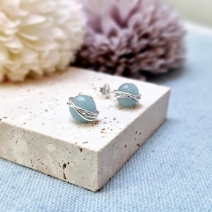 May include: A pair of aquamarine stud earrings. Each earring features a light blue, spherical gemstone wrapped with silver-colored wire. The earrings are displayed on a textured, off-white surface, with blurred floral accents in the background.