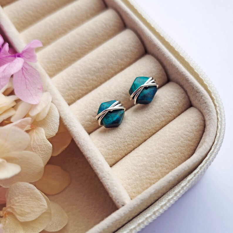 May include: A pair of turquoise gemstone earrings with silver wire wrapping, displayed in a white jewelry box with beige velvet lining.