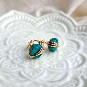 May include: A pair of gold wire wrapped turquoise earrings. The turquoise stones are a deep teal color and have a rough, natural texture.