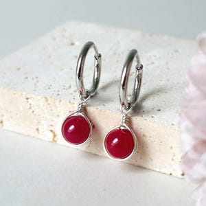 May include: A pair of silver-toned hoop earrings featuring a single round, polished red bead suspended from each hoop. The beads are secured with a delicate silver-toned wire wrap design that encircles the base of each smooth, vibrant red sphere.