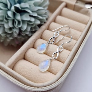 May include: A pair of silver earrings with an infinity symbol design and teardrop-shaped, iridescent moonstone pendants. The earrings are displayed in a beige jewelry box, with a blurred blue-green flower in the background.