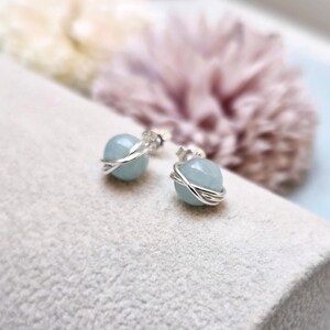 May include: A pair of aquamarine stud earrings. Each earring features a light blue, round gemstone wrapped in a silver wire design. The earrings are displayed on a light gray surface, with a blurred floral background.