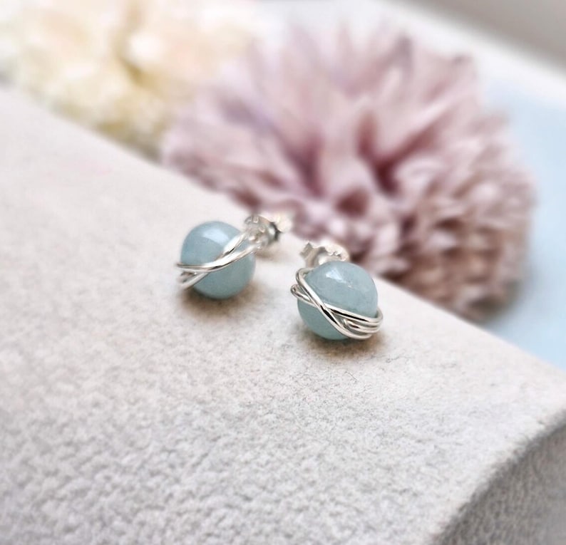May include: A pair of aquamarine stud earrings. Each earring features a light blue, round gemstone wrapped in a silver wire design. The earrings are displayed on a light gray surface, with a blurred floral background.