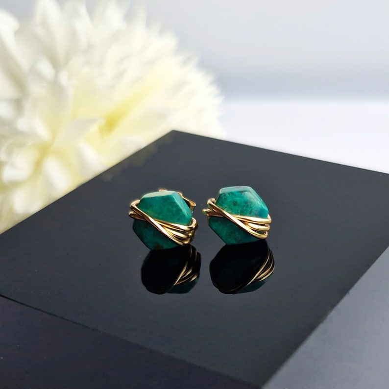 May include: A pair of gold wire wrapped earrings with green gemstone accents. The earrings are on a black surface.