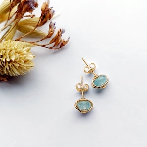 May include: A pair of gold-colored stud earrings with light blue oval-shaped stones. The stones are encased in a wire-like design. The earrings are displayed on a white surface with dried flowers in the background.