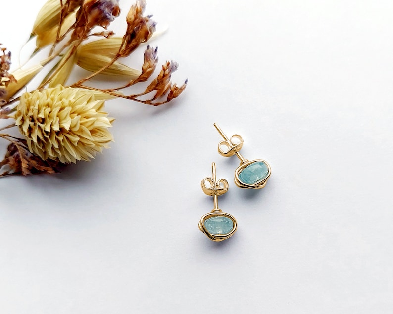 May include: A pair of gold-colored stud earrings with light blue oval-shaped stones. The stones are encased in a wire-like design. The earrings are displayed on a white surface with dried flowers in the background.