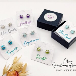 May include: A collection of gemstone stud earrings, including amethyst, clear quartz, aquamarine, turquoise, peridot, and blue opal. Each pair is displayed on a card with the gemstone name. The earrings are in a silver wire setting. A black box with a logo is also present.