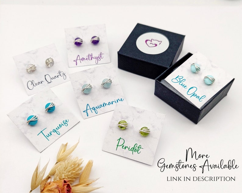 May include: A collection of gemstone stud earrings, including amethyst, clear quartz, aquamarine, turquoise, peridot, and blue opal. Each pair is displayed on a card with the gemstone name. The earrings are in a silver wire setting. A black box with a logo is also present.