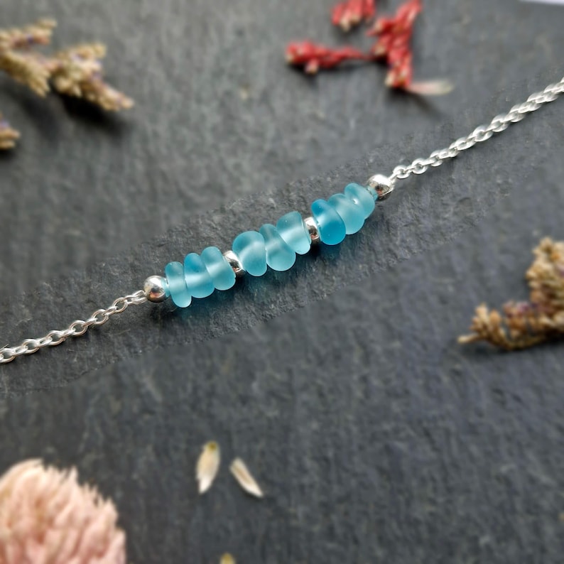 May include: A delicate silver chain necklace with a row of light blue sea glass beads. The necklace is a simple and elegant piece of jewelry.