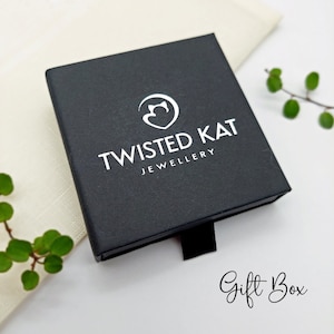 May include: Black gift box with a white logo that says "Twisted Kat Jewellery" and a stylized cat head inside a heart. The box is on a white surface with a green plant in the background.