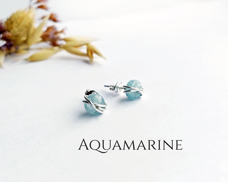 May include: A pair of aquamarine stud earrings. Each earring features a light blue, spherical gemstone wrapped in a silver wire design. The word "AQUAMARINE" is printed in black below the earrings.