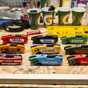 May include: A collection of colorful, personalized multi-tools with names like "LMCA OLD TIMERS", "RFOS", and "Chuck Doyle" printed on them. The tools are red, orange, black, yellow, green, and blue. Other items include Gatorade cups and a headlamp.