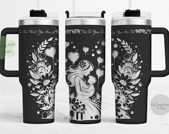 Puede incluir: Una taza de viaje de acero inoxidable negro con un diseño plateado de una madre sosteniendo un bebé, rodeada de flores y corazones. La taza tiene un asa y una tapa con un agujero para pajita. El texto "To the world you are a mother, but to your family you are the world" está grabado en la taza.