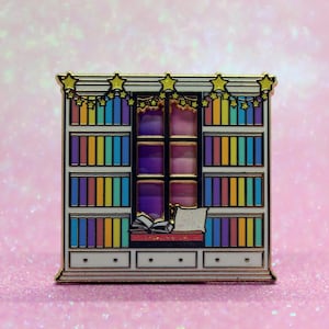 May include: A white bookshelf with a window and a pillow on the bottom shelf. The bookshelf is decorated with a string of yellow stars. The books on the shelves are rainbow colored.