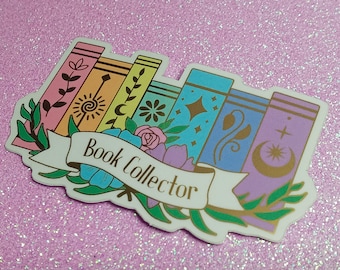 May include: A colorful sticker featuring a stack of stylized books with floral and celestial designs. The books are in shades of pink, yellow, blue, and purple. A banner across the books reads "Book Collector". The sticker is on a glittery pink background.