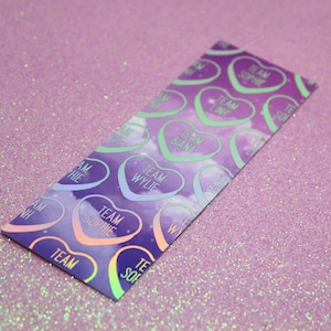 May include: A purple and blue bookmark with a holographic finish. The bookmark features a repeating pattern of hearts with the text "Team Sophie", "Team Lint", "Team Biana", and "Team Wylie" inside each heart.