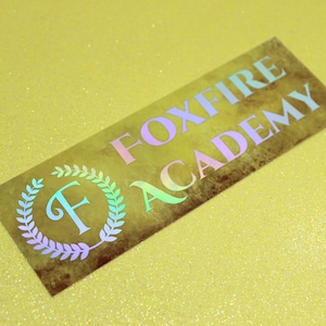 May include: A gold sticker with the text "FOXFIRE ACADEMY" in a rainbow iridescent font. The sticker has a laurel wreath with the letter "F" in the center.